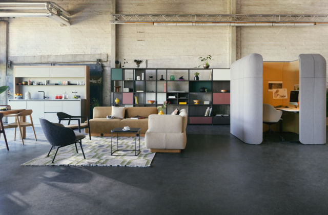 A spacious, industrial-style modern office layout featuring a kitchenette, open shelving, a lounge area with armchairs and sofas on a patterned rug, and a private grey acoustic work pod with a yellow interior.